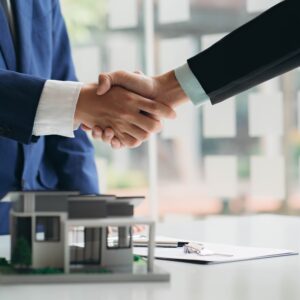 Two business professionals shaking hands over a real estate contract with a house model on the table