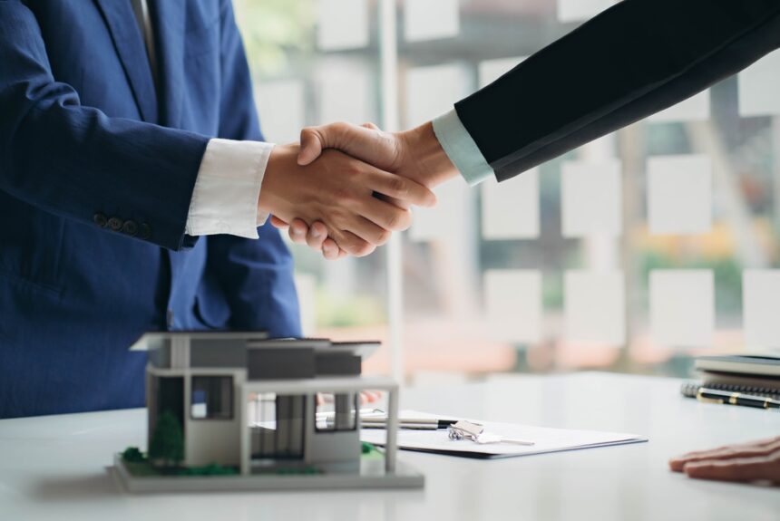 Two business professionals shaking hands over a real estate contract with a house model on the table