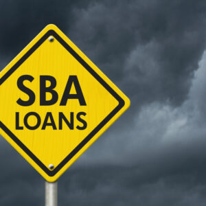 Yellow road sign reading “SBA Loans” with storm clouds in the background, symbolizing upcoming SBA rule changes and challenges in 2025