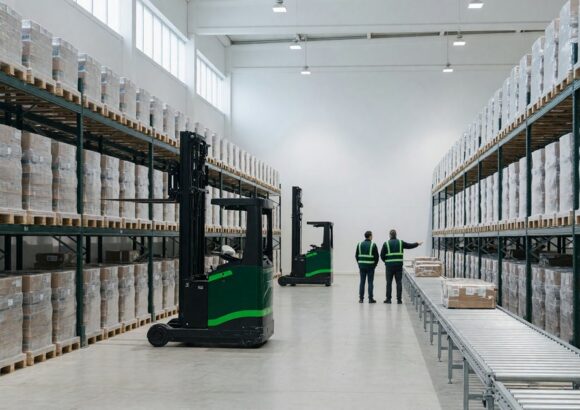 Wide view down the aisle of a modern third-party logistics warehouse: tall deep green pallet racking on both sides loaded with shrink-wrapped pallets, two green reach trucks in the aisle, a roller conveyor running along the right, and two workers in green safety vests seen from behind under bright neutral daylight.