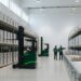 Wide view down the aisle of a modern third-party logistics warehouse: tall deep green pallet racking on both sides loaded with shrink-wrapped pallets, two green reach trucks in the aisle, a roller conveyor running along the right, and two workers in green safety vests seen from behind under bright neutral daylight.