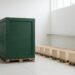 A business owner stands with their back to the camera in a bright warehouse, looking at one oversized deep green crate that towers over a row of much smaller crates, a visual analogy for one customer carrying most of the revenue.